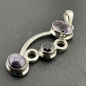 Puede incluir: Un colgante de plata con tres piedras preciosas ovaladas. Las piedras preciosas son de color morado, rojo y azul. El colgante está diseñado en un estilo moderno con una forma curva y tres círculos.