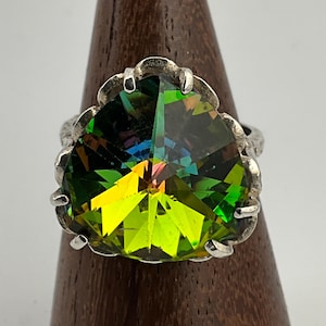 May include: A silver ring featuring a large, faceted gemstone with a vibrant display of colors, including green, yellow, and hints of blue and pink. The gemstone is set in a textured silver band with a unique, scalloped edge.