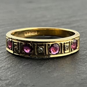 May include: A yellow gold band ring featuring a series of alternating round faceted deep pink gemstones and small clear white stones set within rectangular segments, with a milgrain edge detail along the top and bottom of the band.
