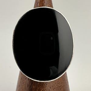 Puede incluir: Un anillo ovalado con un centro negro brillante y un borde plateado. El anillo se exhibe sobre un soporte de madera. El centro negro tiene un diseño sutil en forma de cuadrado.