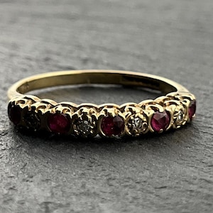 May include: A gold ring with alternating red gemstones and small clear stones. The ring has an intricate, ornate design, with the gemstones set in individual bezels. The ring is shown against a dark, textured background.