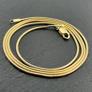 Op de afbeelding: Een goudkleurige slangenketting met een glad, flexibel en afgerond schakelontwerp. De ketting is in een cirkelvormig patroon gewikkeld en bevat een veilige karabijnsluiting aan één uiteinde, wat zorgt voor een gepolijste en uniforme uitstraling.