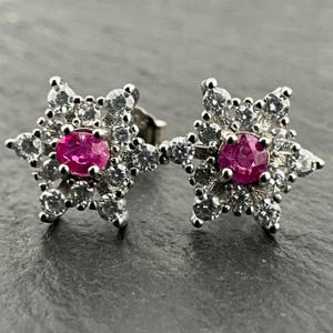 Könnte beinhalten: Ein Paar sternförmige Ohrstecker. Jeder Ohrring hat einen leuchtend rosa Edelstein in der Mitte, umgeben von kleineren, klaren Edelsteinen. Die Ohrringe sind in einem silberfarbenen Metall gefasst und liegen auf einer dunkelgrauen Oberfläche.