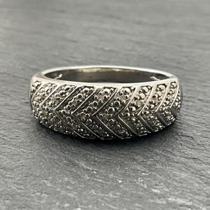 May include: A silver ring with a chevron pattern. The ring is made of a shiny metal and features small, round embellishments creating a textured surface. The design is a series of angled lines forming a repeating V-shape.
