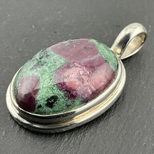 May include: A silver pendant featuring a green and red gemstone. The gemstone is oval shaped and has a smooth, polished finish.