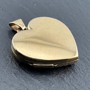 May include: A heart-shaped gold locket pendant with a smooth, polished surface and a hinged bail at the top. The metal shows fine surface scratches consistent with wear, and the locket is closed, displaying a solid, plain gold exterior finish.