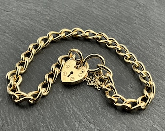 Op de afbeelding: Een goudkleurige kettingarmband met een hartvormige hangslot sluiting. De armband heeft een reeks in elkaar grijpende schakels. Het hangslot heeft een sleutelgat en is gegraveerd met kleine details. Een kleine bedel hangt aan de sluiting.