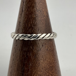 May include: A silver-colored ring with a twisted rope design. The ring is displayed on a wooden cone-shaped stand. The ring's texture and design are clearly visible, showcasing its intricate details. The ring is a piece of jewelry.