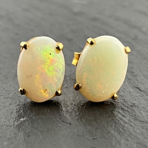 May include: A pair of oval-shaped stud earrings featuring milky white opals with iridescent flashes of green and orange color play. Each opal is securely held in a simple four-prong yellow gold setting, showcasing a smooth, polished surface.