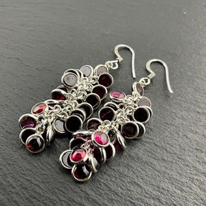 Pode incluir: Brincos pendurados em prata com aglomerados de pequenas pedras preciosas redondas, vermelho escuro e roxo. Os brincos têm fecho de gancho e são exibidos em uma superfície cinza escuro.
