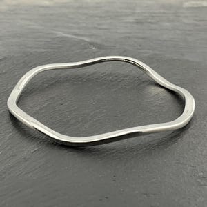May include: A silver-coloured, wavy bangle bracelet. The bracelet has an irregular, organic shape, with smooth, polished surfaces. The design is minimalist and modern. The bracelet is approximately 6.35 cm in diameter.