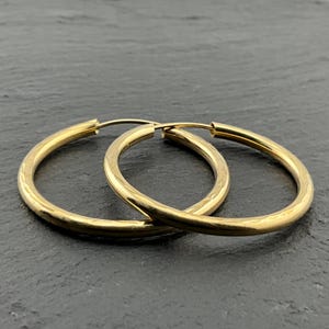 Op de afbeelding: Een paar gepolijste gouden oorringen met een glad, afgerond profiel. De oorbellen hebben een klassiek rond ontwerp met een dunne draadsluiting en een reflecterende metalen afwerking die het licht op het gebogen oppervlak opvangt.