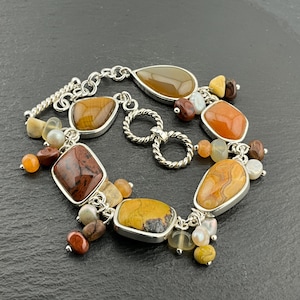 May include: A silver bracelet featuring a variety of colorful, irregularly shaped gemstones. The bracelet includes stones in shades of brown, orange, yellow, and cream, with a mix of textures and shapes. The bracelet is on a dark surface.