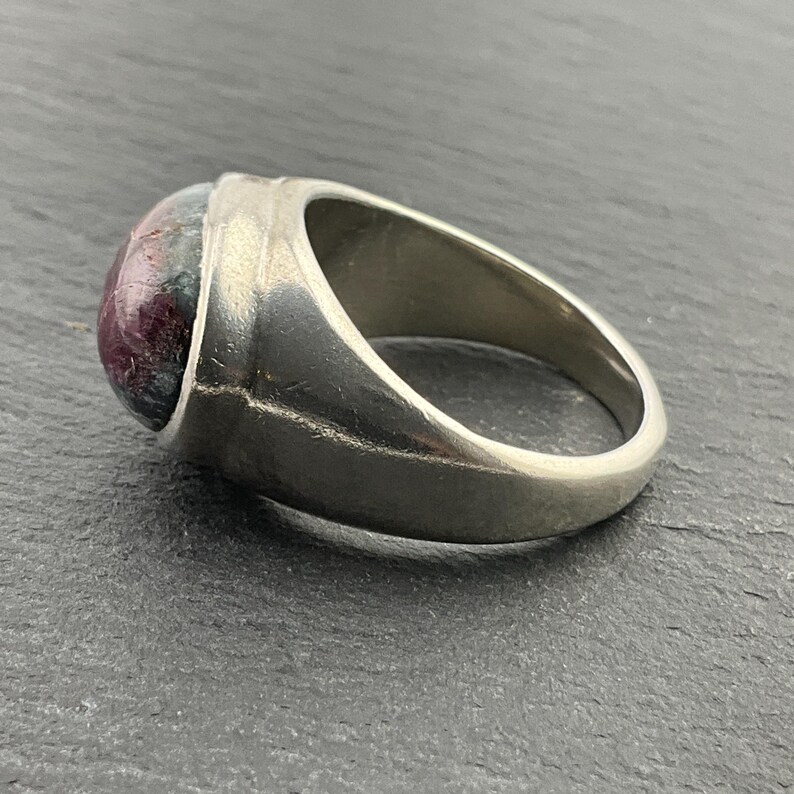 May include: A silver ring with a large, round, cabochon-cut gemstone. The gemstone is a deep red color with green and black inclusions.