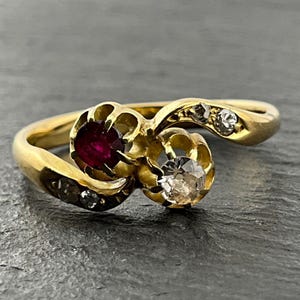 May include: A gold bypass ring featuring a central round red gemstone and a round clear gemstone, each set in a scalloped gold setting. The band is accented with two small clear stones on each side, creating a continuous flowing design.