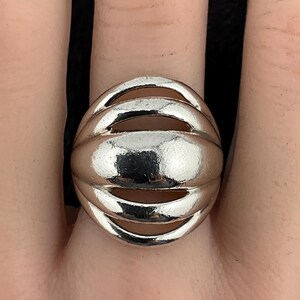 Pode incluir: Um anel prateado com um design arredondado e canelado. O anel tem uma série de ranhuras horizontais que criam uma superfície texturizada. O anel é usado em um dedo e o metal reflete a luz, criando destaques e sombras.