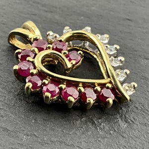 May include: A gold heart-shaped pendant featuring a row of round, deep red rubies set along the inner curve and a row of small, clear, round-cut diamonds set along the outer edge, complete with a gold bail at the top for a necklace chain.
