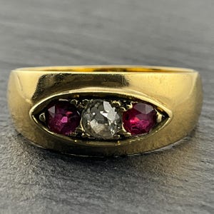 Pode incluir: Um anel de ouro com uma fila horizontal de três pedras cravadas numa moldura oval rebaixada. A pedra central é um diamante transparente facetado, ladeado de cada lado por um rubi redondo vermelho escuro. O aro tem um acabamento polido.