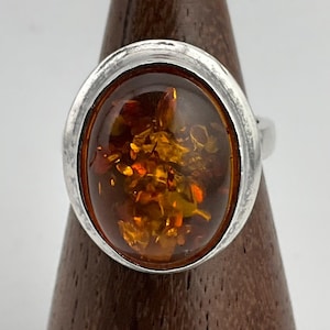 May include: Silver ring with an oval amber stone. The amber has golden inclusions and is set in a textured silver bezel. The ring is displayed on a wooden stand. The amber stone is approximately 2.5 cm in length.