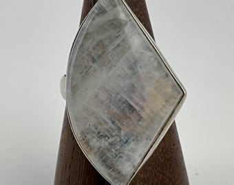 May include: A large, silver-toned ring featuring a faceted, iridescent, rectangular gemstone. The gemstone has a pearlescent, translucent appearance with internal veining and color variations. The ring is displayed on a wooden stand against a white background.