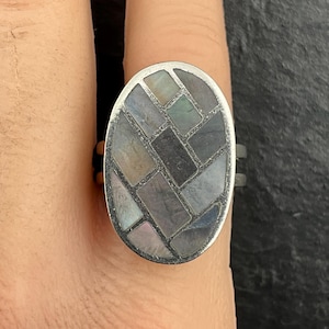 Könnte beinhalten: Silberring mit ovaler Fassung. Die Fassung ist mit einem Mosaikmuster aus rechteckigen Teilen in Grau-, Blau- und schillernden Farbtönen eingelegt. Der Ring befindet sich an einem Finger.