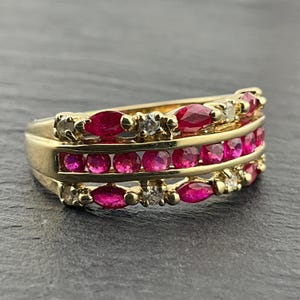 May include: A gold ring featuring three horizontal rows of gemstones. The top and bottom rows contain alternating marquise-cut red rubies and small round white diamonds. The middle row consists of a continuous line of round red rubies.