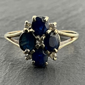 May include: A gold ring featuring a cluster of four oval-cut deep blue sapphires arranged in a cross pattern, accented by small round clear stones set between the sapphires. The band is thin and splits into two sections as it reaches the setting.