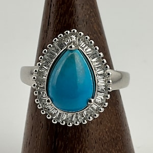 May include: A silver ring with a large, teardrop-shaped, bright turquoise stone. The stone is encircled by a halo of small, rectangular, clear stones. The ring is displayed on a wooden stand.
