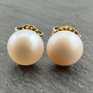May include: A pair of round, light peach-colored pearl stud earrings. The pearls have a subtle luster and are set on gold-colored posts with decorative detailing. The earrings are displayed on a dark gray surface.