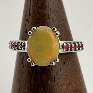May include: A silver ring with a large oval opal gemstone surrounded by a halo of small red rubies. The ring is set on a wooden ring holder.