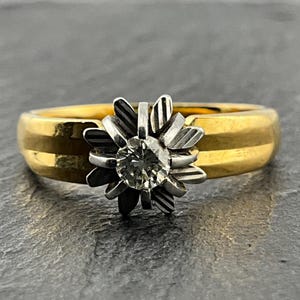 May include: A gold ring featuring a central round diamond set within a silver-toned, multi-petaled flower-shaped mount. The band is smooth and polished yellow gold, while the petals surrounding the diamond have a textured, striped finish.