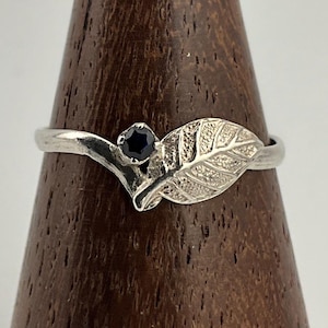 May include: A silver ring with a leaf motif and a dark blue gemstone. The leaf design has detailed textures. The ring is presented on a wooden stand, highlighting its delicate design and nature-inspired theme. The ring's design is reminiscent of a leaf.