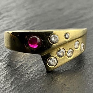 Puede incluir: Un anillo de oro con diseño cruzado que presenta una piedra preciosa roja redonda en un lado y siete pequeñas piedras claras redondas incrustadas en el otro, con una superficie metálica pulida y altamente reflectante.