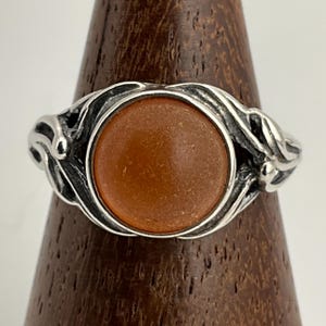 May include: A silver ring featuring an oval, amber-coloured stone set within a decorative, swirling design. The ring's band has an intricate, leaf-like pattern, giving it a vintage or antique look. The stone is a warm, earthy tone, contrasting with the silver.