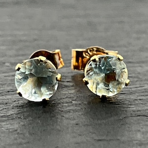 May include: A pair of stud earrings featuring round, faceted, pale blue gemstones set in gold metal settings. Each earring has a circular stone with multiple facets that catch the light, secured by a gold post and backing mechanism.