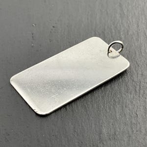 May include: A rectangular, silver pendant with a small loop at the top. The pendant has a smooth, reflective surface and rounded corners. The jewellery piece is likely made of metal and is suitable for engraving.