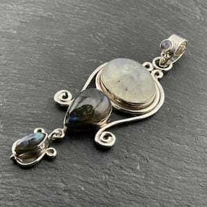 May include: A silver pendant with three gemstones: a large oval moonstone, a teardrop-shaped labradorite, and a small oval labradorite. The pendant has a decorative, swirling design.