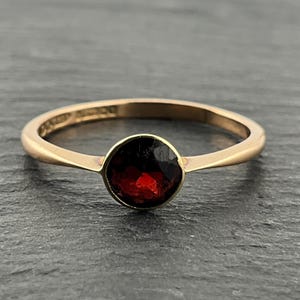 May include: A gold ring featuring a single, round, faceted deep red garnet gemstone set in a smooth bezel setting. The band is thin and polished, with a simple, continuous design that tapers slightly as it meets the central stone setting.