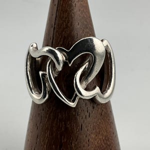 May include: A silver ring with a heart-shaped design. The ring features three interconnected hearts, creating a continuous loop. The polished metal reflects light, highlighting the curves and details of the design. The ring is displayed on a wooden stand.