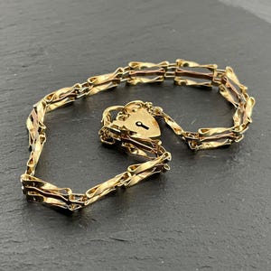 Puede incluir: Una pulsera de tono dorado que presenta una serie de eslabones rectangulares retorcidos e interconectados. La pulsera incluye un dije de candado en forma de corazón con un corte de cerradura visible, unido a la cadena con una argolla.