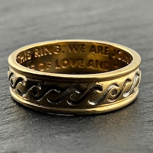 May include: A gold-toned metal ring featuring a continuous embossed wave pattern around the exterior band. The interior of the ring is engraved with the visible text: THIS RING. WE ARE JOINED IN A STATE OF LOVE AND PEACE.