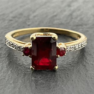 May include: A gold ring featuring a large emerald-cut deep red gemstone at the centre, flanked by two smaller round red side stones and accented with a row of tiny clear white stones along the band on both sides.