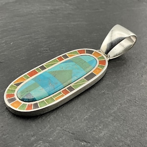May include: A silver pendant with an oval turquoise stone inlay. The stone is a mosaic of blue and green squares. The pendant has a colourful border of orange, red, black, and green. The pendant is approximately 5 cm long.