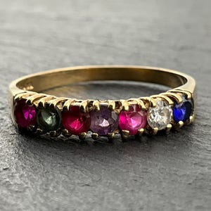 Op de afbeelding: Een gouden ring met een rij van zeven rond geslepen edelstenen in verschillende kleuren, waaronder rood, groen, paars, wit en blauw. De stenen zijn gezet in een klauwzetting aan de voorzijde van de gepolijste geelgouden ringband.