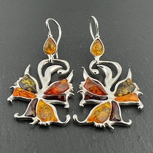 Pode incluir: Um par de brincos pendurados prateados com um design floral. Cada brinco tem uma pedra de âmbar em forma de gota na parte superior e quatro pétalas de cor âmbar. As pedras de âmbar variam em cor de amarelo claro a castanho escuro.