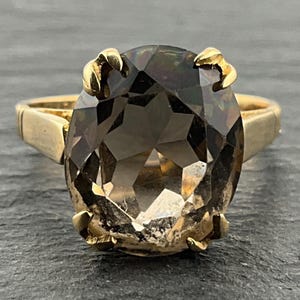 Könnte beinhalten: Ein Goldring mit einem großen, oval geschliffenen, durchscheinenden dunkelbraunen Edelstein, der von vier markanten Krappen gehalten wird. Die Ringschiene ist glatt und poliert und unterstreicht das schlichte Design des facettierten Steins.