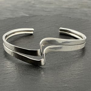 May include: A silver cuff bracelet with a modern, minimalist design. The bracelet features two parallel bands that curve and overlap, creating a wave-like shape. The polished metal reflects light, highlighting the smooth, sleek lines.