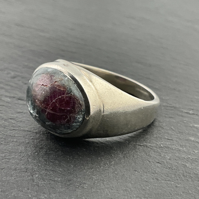 May include: A silver ring with a large, oval-shaped, red gemstone set in the center. The gemstone has a unique, textured surface.