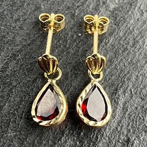 May include: A pair of gold drop earrings featuring a teardrop-shaped dark red gemstone set in a textured gold frame. Each earring has a small gold shell-like accent above the stone and a thin gold post with a matching gold butterfly back.