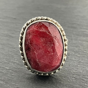 May include: A silver ring featuring a large, oval-shaped, red gemstone set in a twisted rope design.